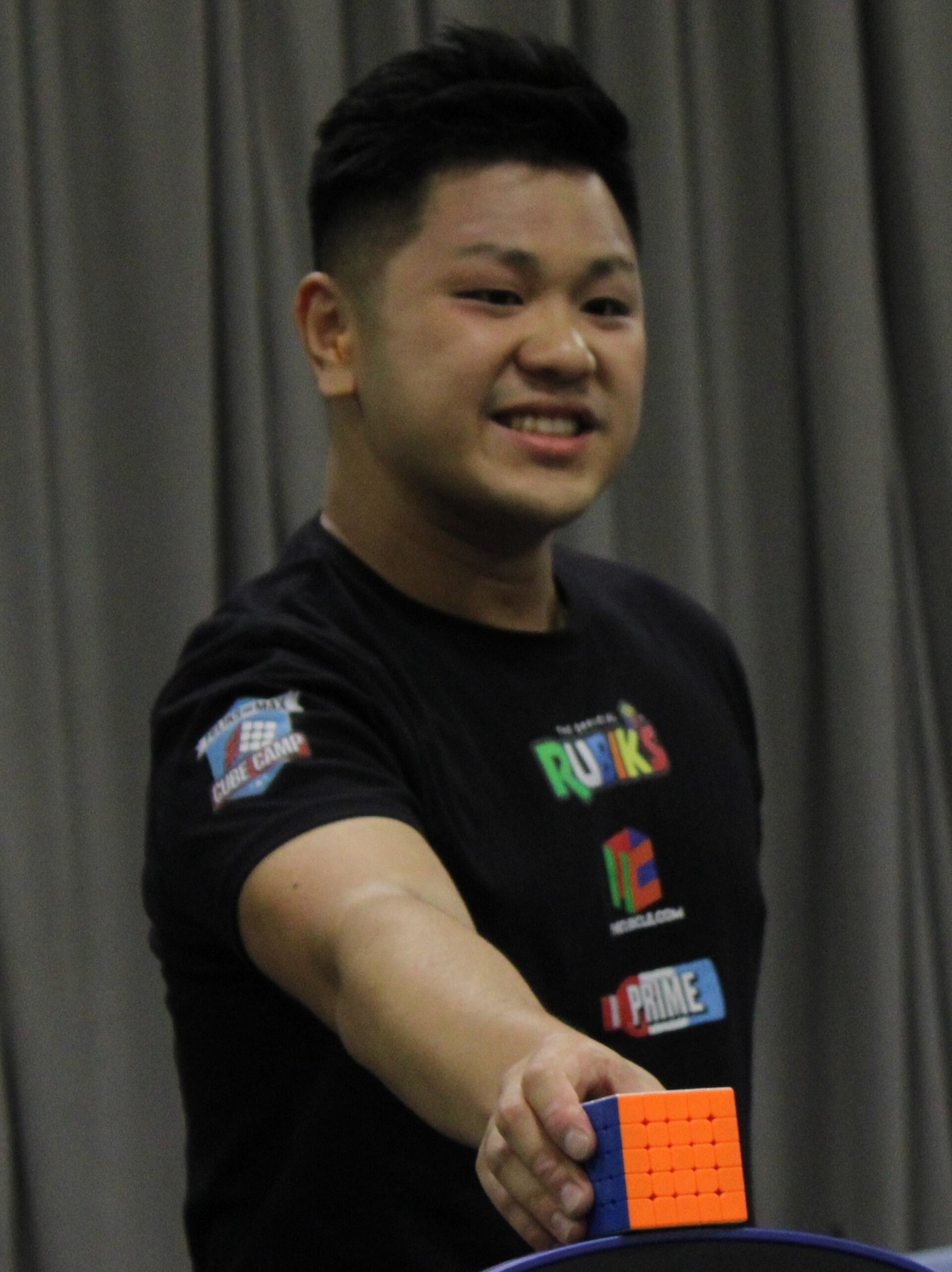 Max Park sonríe con el brazo extendido y apoya sobre el cronómetro un cubo de 5x5 recién resuelto, con remera negra y telón gris de fondo.