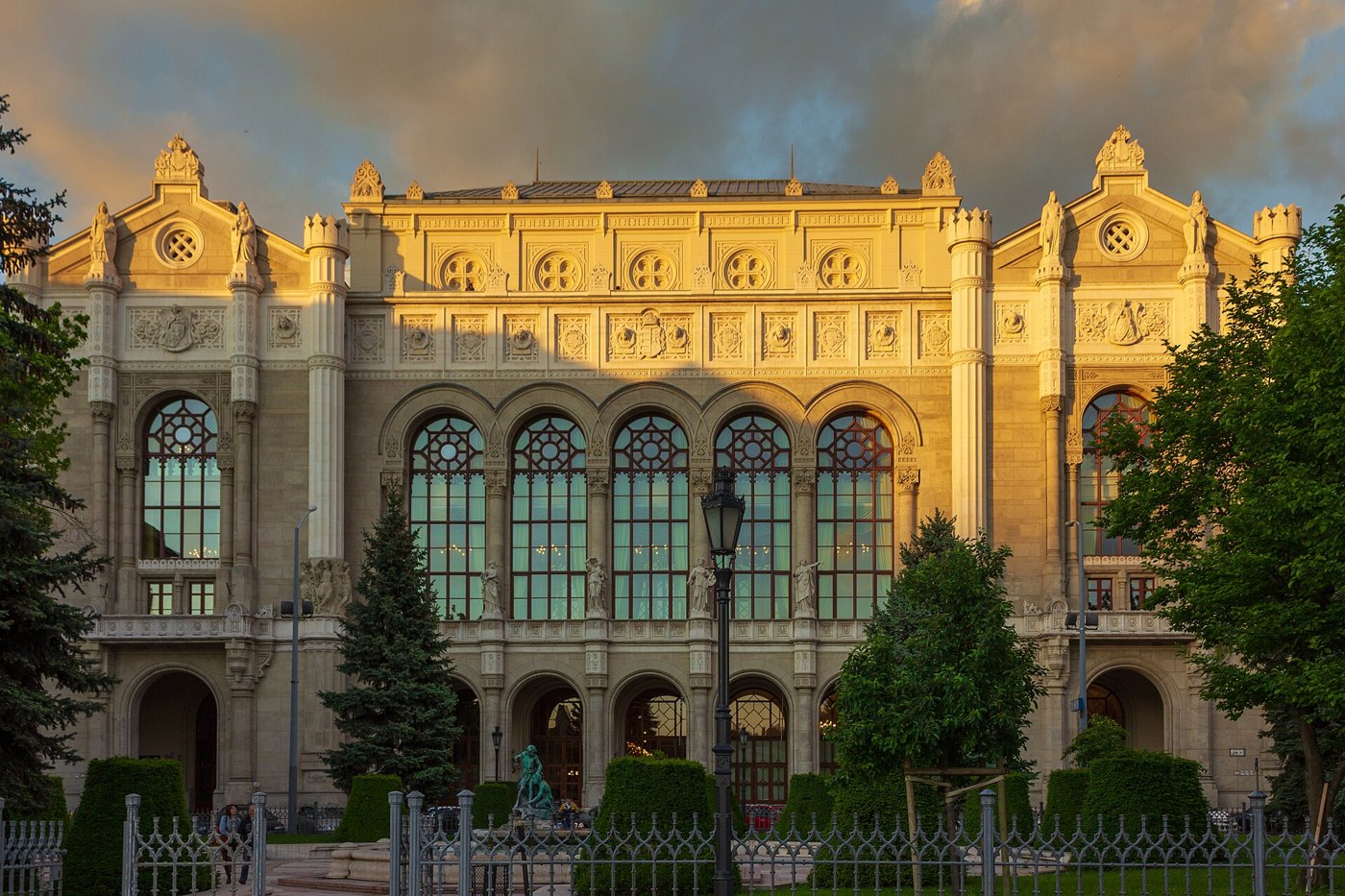 Fachada del edificio Vigadó de Pest, en Budapest, con ventanales en arco y árboles en primer plano, iluminada por la luz del atardecer.