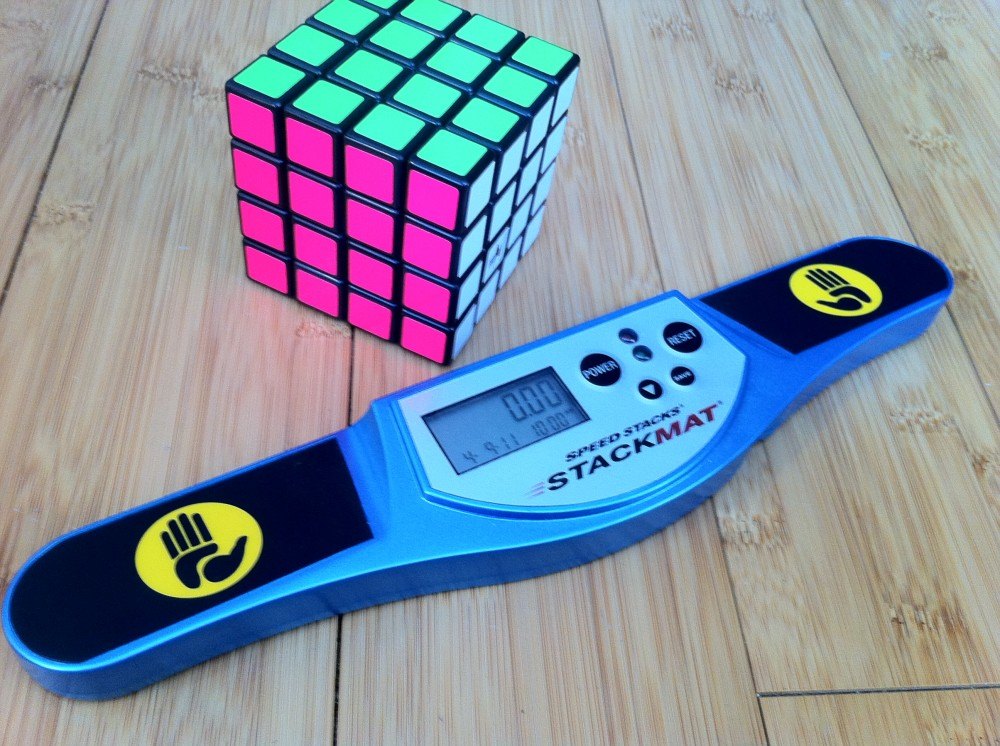 Cubo de 4x4 con caras rosa y verde junto a un cronómetro Stackmat azul con dos sensores amarillos, sobre una mesa de madera clara.